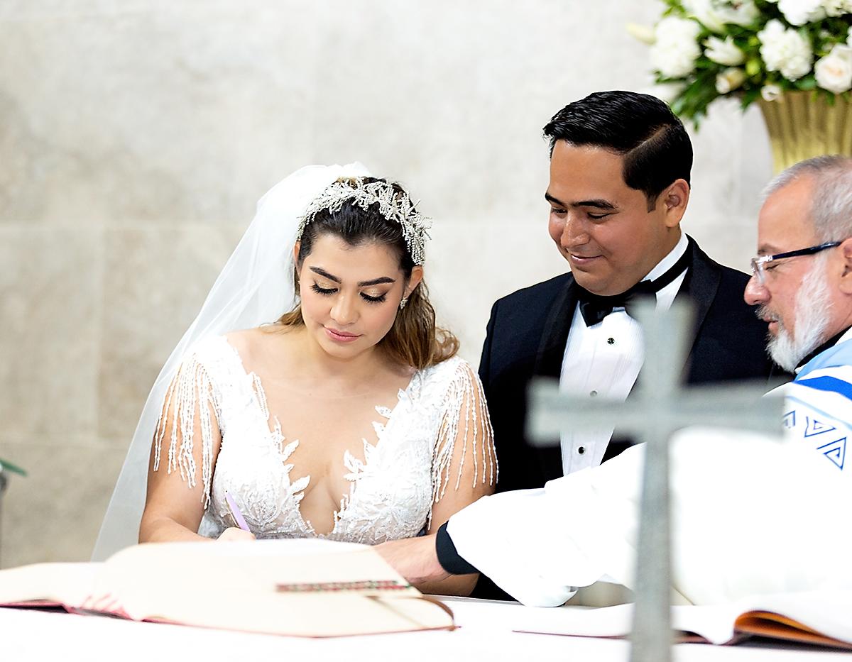 La novia, vestida en un diseño de escote en V y tocado de pedrería, firma el registro de matrimonio ante la vista de su esposo.