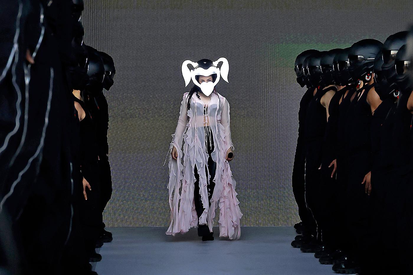 Rosalía entró al escenario con unos pantalones y bustier negros, un sobretodo color rosa de la marca sueca Acne, gafas futurísticas de Rick Owens y los cascos de sus motopapis, diseño del español Carlos Sáez.