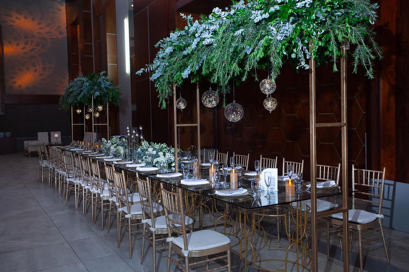 Mesas estilo imperial de vidrio decoradas con arreglos en diferentes niveles con detalles en metal, transparencias, velas eternas, flores y follaje.