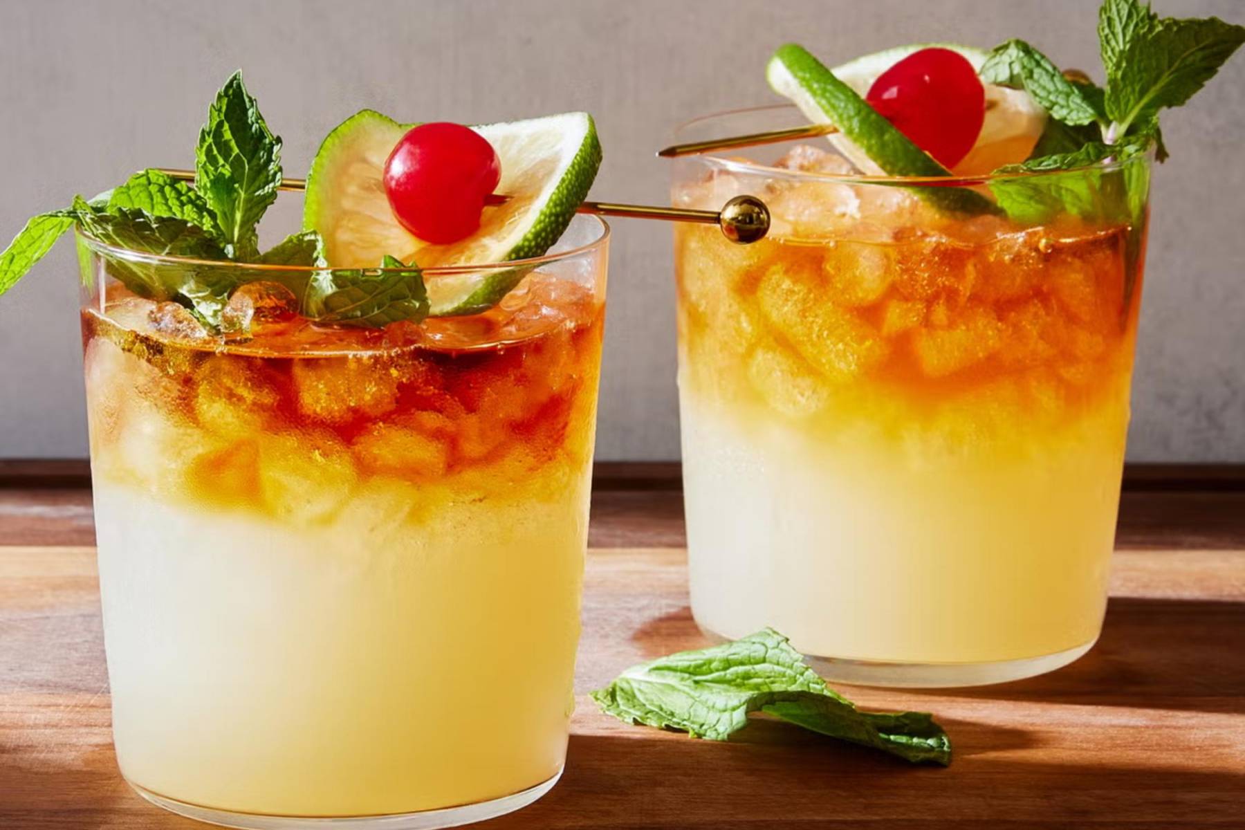 <b>Mai Tai:</b>Nacido después de la Segunda Guerra Mundial, es el cóctel precursor de los famosos tiki cocktails, inspirados en la cultura polinesia. Su nombre proviene de la expresión tahitiana <i>mai tai</i>, que significa “delicioso”, y se ha convertido en un clásico entre las bebidas tropicales. Esta mezcla exótica combina ron jamaicano, ron añejo, Cointreau, amaretto, zumo de piña, zumo de lima y granadina. Para prepararlo, mezcla 30 ml de ron jamaicano, 30 ml de ron añejo, 15 ml de Cointreau, 15 ml de amaretto, 30 ml de zumo de piña, 10 ml de zumo de lima y 10 ml de granadina en una coctelera con hielo. Agita bien y sirve en un vaso corto con hielo, decorado con hierbabuena, una rodaja de lima con una cereza. ¡Una explosión tropical en cada sorbo!