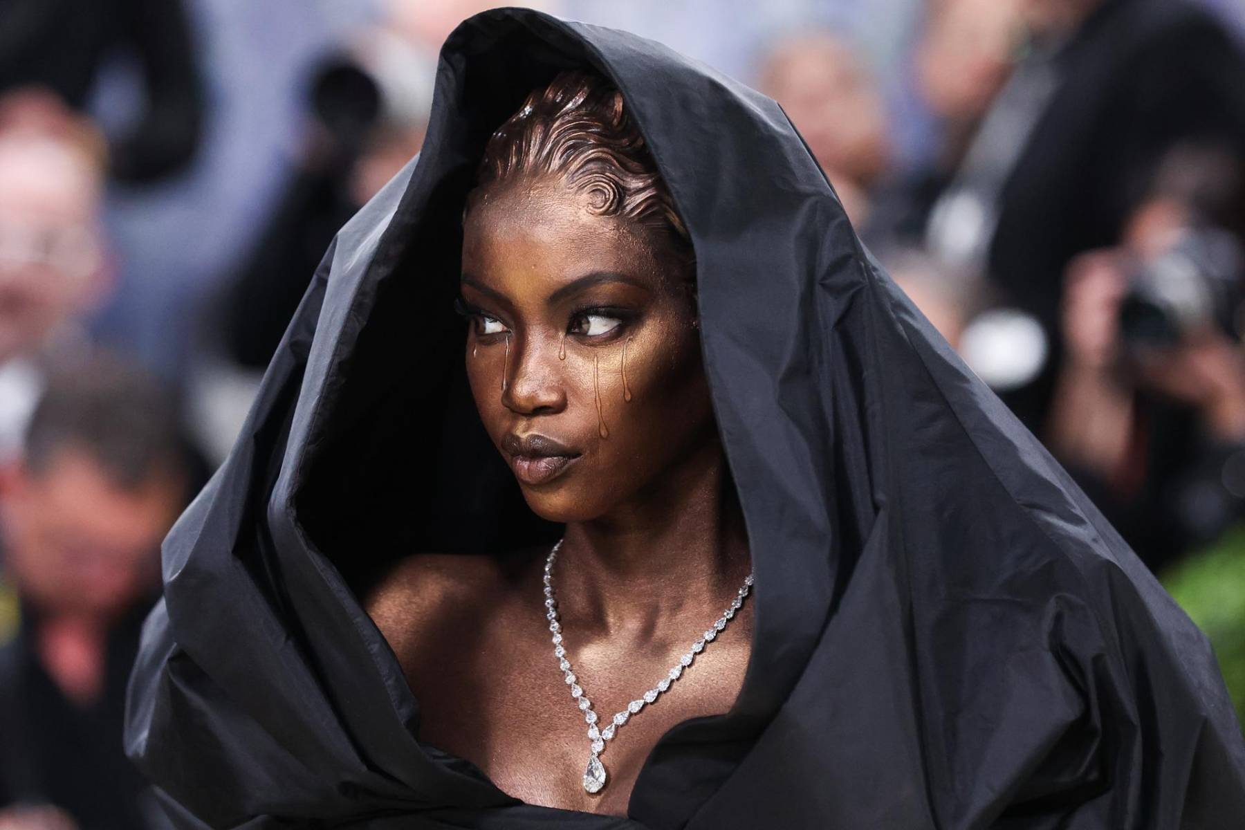 Anok Yai impactó con su escultórico look complementado con una capucha negra profunda que enmarcaba su rostro, evocando una estatua andante o una "madonna negra". El aspecto más comentado fue su maquillaje teatral, que simulaba lágrimas inmovilizadas o bronce derretido cayendo por sus ojos, creado para representar el dolor y la esperanza.