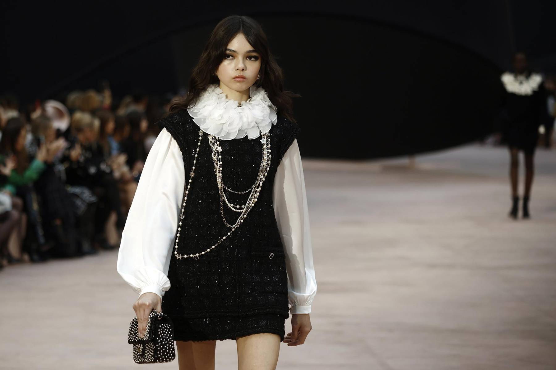 Cuellos con volantes desmontables que se asemejan a corolas, gabardinas de encaje de lana negra con incrustaciones de piel sintética realzadas y suéteres negros con plastrones de pétalos blancos en 3D marcan la colección con un toque poético.