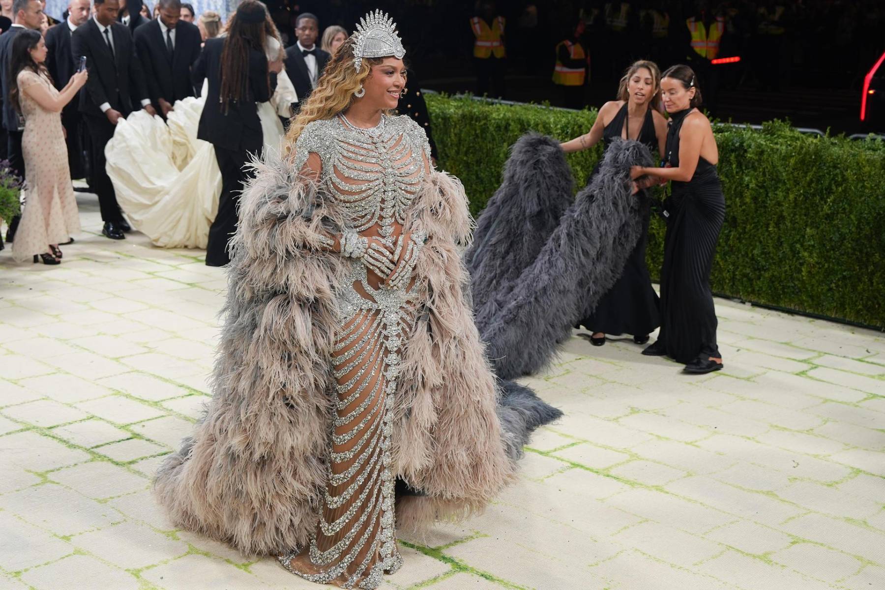 Beyonce regresó a la Met Gala con un diseño de Olivier Rousteing cubierto de cristales que dibujaban un esqueleto, jugando directamente con la idea de “Fashion is Art” y la anatomía como lienzo. La pieza, casi etérea, se completó con una capa de plumas de gran volumen, joyería de alto impacto —incluida una corona— y una melena en ondas doradas que suavizaba la fuerza del look