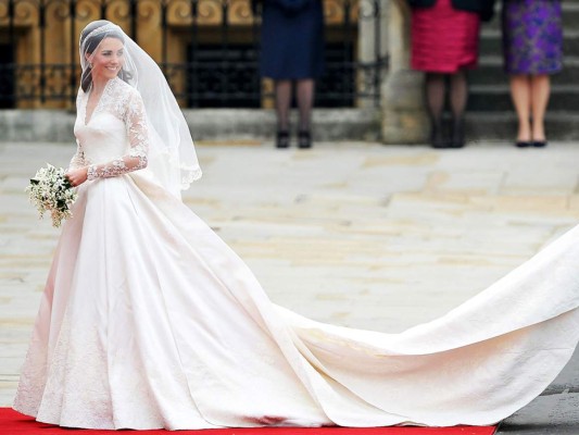 Kate Middleton. La discreta prometida del príncipe William deslumbró con su memorable diseño color marfil y blanco satinado con escote corazón realizado por Sarah Burton para Alexander McQueen. Ese 28 de abril de 2011 Kate lucía espectacular con su silueta envuelta en encaje francés y una voluminosa falda con una cola de tres metros de largo con aplicaciones de encaje y flores de seda. El corpiño de satén fue inspirado en la tradición victoriana de corsetería -estrecho en la cintura y acolchado en las caderas- detalle que caracteriza los diseños de Alexander McQueen. En la espalda, acabada en gazar, destacaban botones forrados de organza sujetados por lazos. El velo, elaborado en capas de tul suave de seda color marfil con recorte de flores bordadas a mano por la Real Escuela de Costura, se complementó con la tiara Cartier que Isabel II recibió como regalo de su madre por su 18 cumpleaños. Kate llevó además pendientes de diamantes en forma de pera que la firma Robinson Pelham diseñó inspirándose en el escudo de armas de la familia Middleton y sus zapatos fueron también hechos a mano por el equipo de Alexander McQueen. Finalmente el ramo, diseñado por Shane Connolly, fue inspirado en las flores favoritas de la Familia Real británica y la familia Middleton e incluía lirios, jacintos, hiedras y mirto.