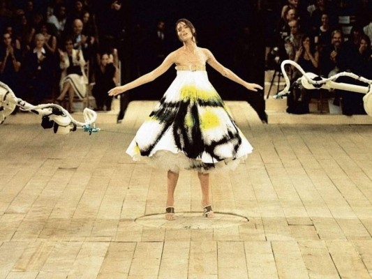 Alexander McQueen<br/>Si eres aficionada de la moda, es imposible que olvides este momento en la pasarela de 1998, del diseñador Alexander McQueen. Para el cierre del show, Shalom Harlow modeló un simple vestido blanco, cuando en cuestión de segundos se convirtió en una obra de arte, al ser rociada con pintura por unos robots.<br/>