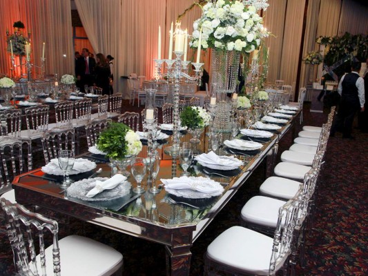 Mesas estilo imperial con sillas acrilícas, candelabros de cristal, bases de vidrio con rosas, margaritas y hortensias.