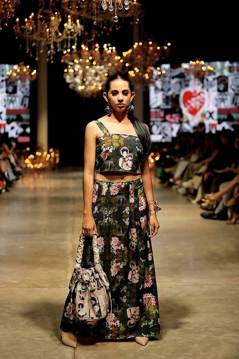Este conjunto floral combina un crop top de tirantes con una falda larga, ambos en tonos oscuros y estampados coloridos que evocan motivos religiosos y naturales. La bucket bag da un contraste al look.