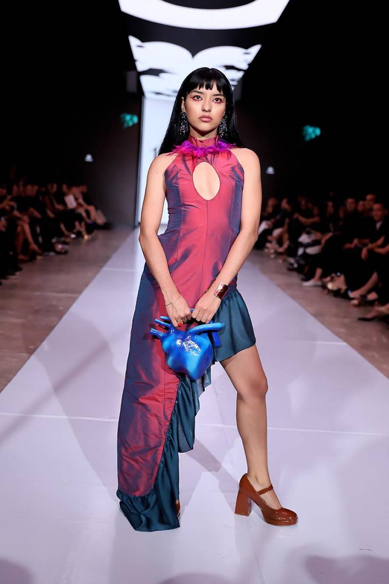 Vestido tornasol en rojo y azul que juega con la asimetría y los cortes cut out, evocando la apertura ritual del pecho. El cuello halter adornado con plumas en tonos rosados y violetas se enlaza con un escote profundo en la espalda, mientras los aretes representan las noches estrelladas de los antiguos estudios mayas; y cartera en forma de corazón azul, símbolo de un alma extraída y ofrecida en el eterno ciclo de creación y destrucción.