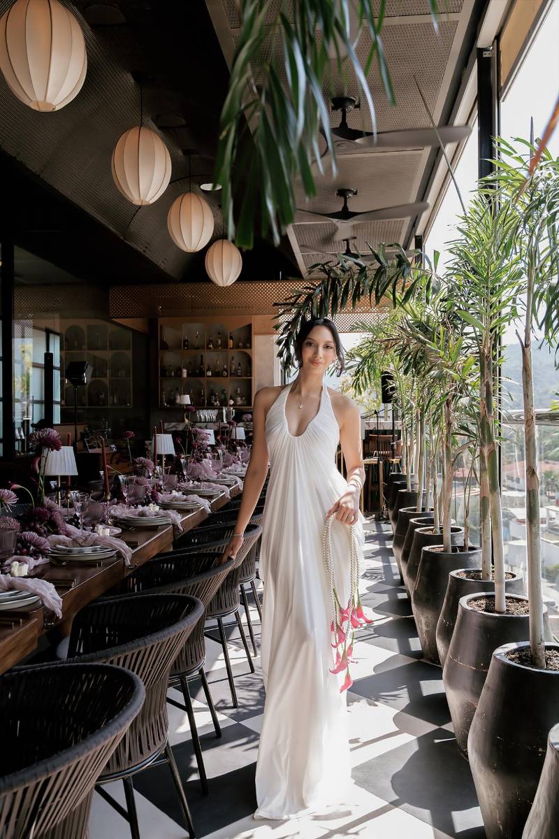 La novia lució un vestido marfil de silueta fluida, con escote halter profundo y delicados pliegues desde el cuello que crean una caída ligera y etérea hasta el suelo.