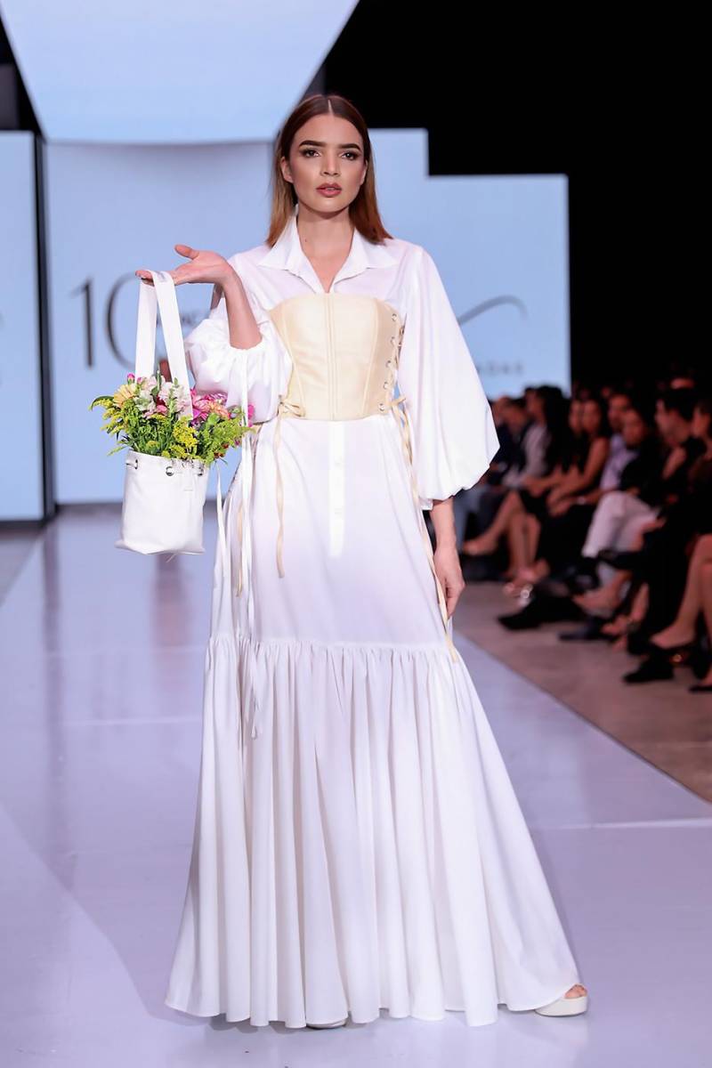 Conjunto de dos piezas compuesto por un faldón en algodón egipcio blanco, con cuello clásico, botonadura frontal, mangas abullonadas y falda fruncida que aporta volumen y movimiento. Se complementa con un corsé en shantung de seda beige, adornado con lazos largos que también se repiten en las mangas. Como accesorio, destaca una cartera decorada con flores naturales, aportando frescura y un toque artesanal