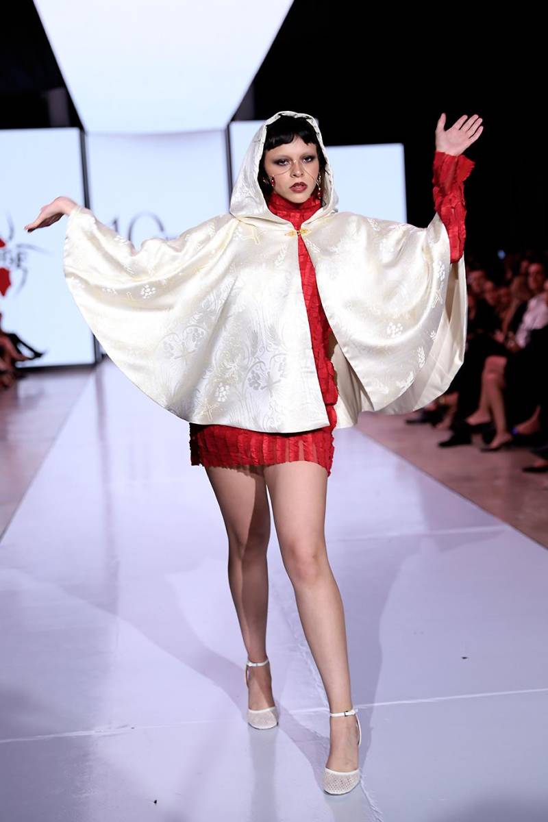 La modelo Fabiana Urcina abrió la pasarela con un impactante vestido de mesh rojo, cuello alto y asimetría en mangas, adornado con vuelos bordados. Completó el look una capa con capucha en satín ivory con brocado dorado y un cut-out en forma de cruz en la espalda.