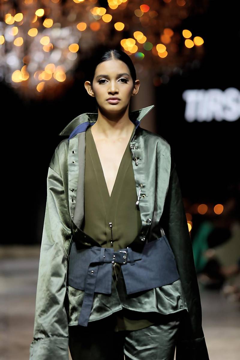 El layering se presenta de manera audaz combinando blusa y long jacket de ojales metálicos con imperdibles oversize. Dibujando la silueta, un cinturón de tela superpuesto en tono contrastante.