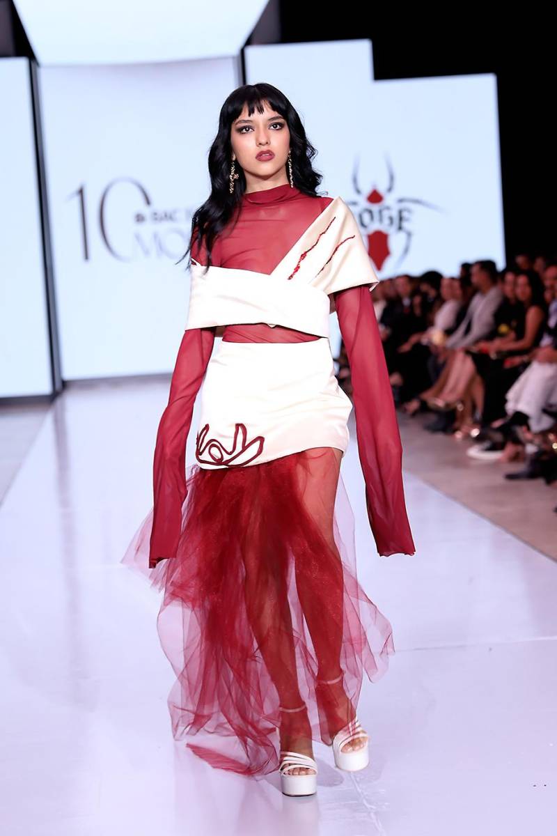 Blusa de mesh rojo con mangas extra largas bajo una estola drapeada de podesua ivory con bordado de piedras rojas. La falda, también en podesua ivory, lució tiras de chiffon y tul rojo que aportaron movimiento y dramatismo.
