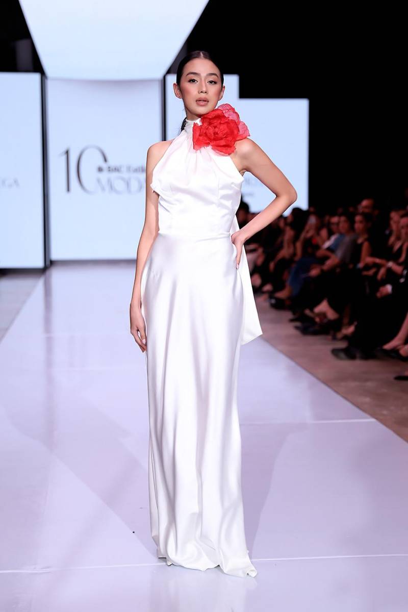 Vestido blanco corte en ‘A’ con escote halter y flor roja sobredimensionada en el cuello, simbolizando la verdad que rompe la inocencia y el primer destello de amor en la historia de Blanca Olmedo