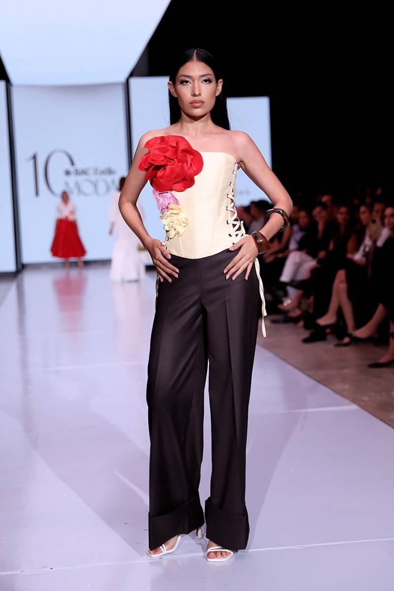 Conjunto de dos piezas conformado por un corsé en shantung de seda color beige, decorado con delicadas flores del mismo textil en distintos tonos. El diseño incorpora ojetes laterales con largas tiras que permiten ajustar la silueta. Se complementa con un pantalón de cashemere en tono café y ruedo de acabado estilo valenciano.