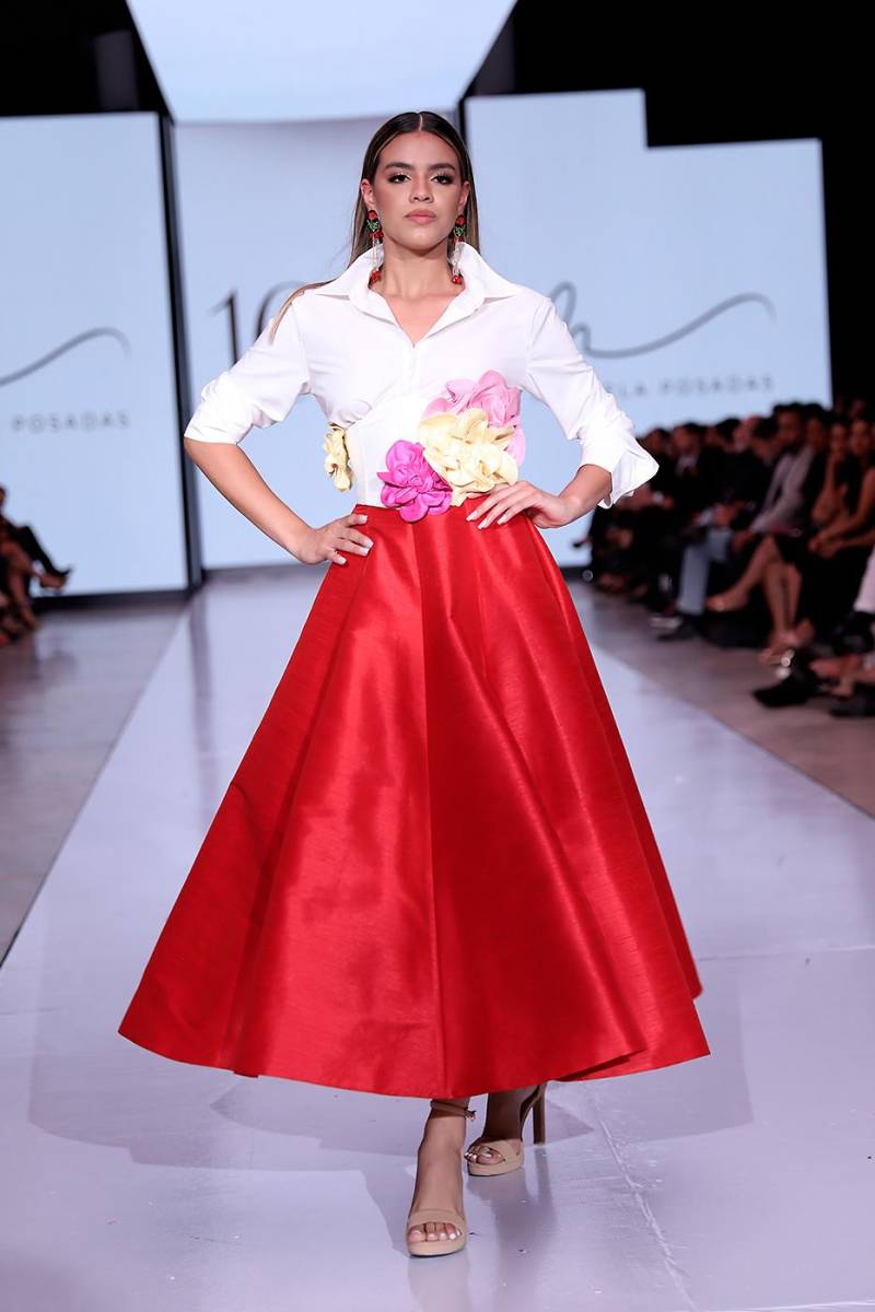 Conjunto de tres piezas compuesto por una camisa clásica de botones en algodón egipcio blanco, acompañada de un corsé en shantung de seda blanco, decorado con flores en diversos colores. El look se completa con una falda estilo “A” en shantung de seda rojo, estructurada con pliegues tipo cuchilla