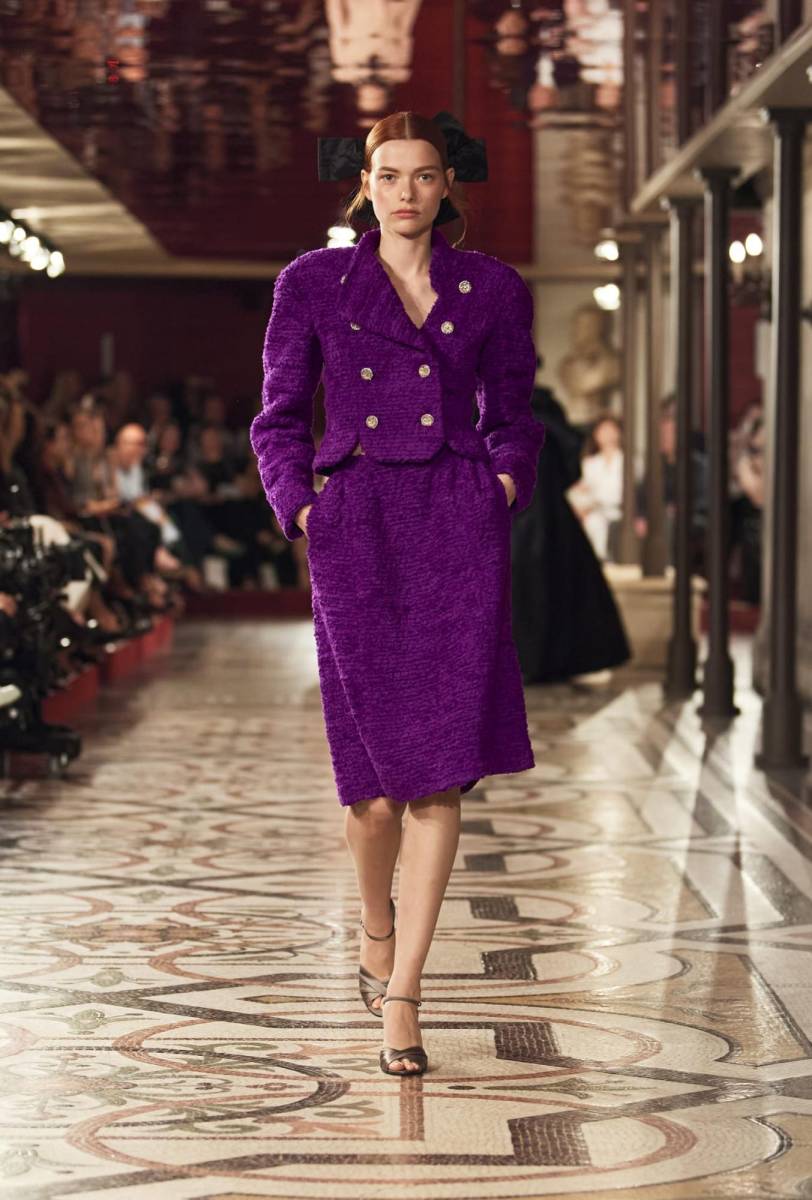 Trajes de tweed revisitados, un esmoquin de pana negro y una blusa blanca con plastrón bordado, un traje negro con un largo culotte y chaqueta corta y entallada con hombros envueltos en plumas negras, bailan entre abrigos largos, capas voluminosas y vestidos de noche, que evocan una tradición escénica modernizada.